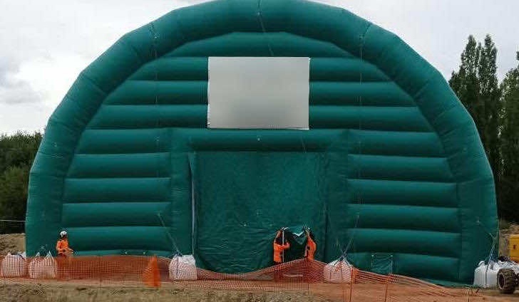 View of the front facade, the front gable of a very large inflatable shed module in the shape of a half-moon tunnel, with a large main access door. Two technicians are finalizing the positioning of the main access door. This inflatable structure is made of 0.45 mm double-skin padded PVC, very strong and very robust, very versatile too.