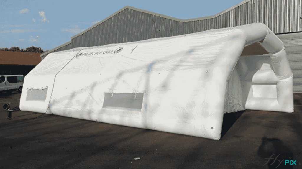 A general view of the inflatable first aid shelter, with a captive air system and usable internal dimensions of L = 12 m x W = 6 m, that we manufactured for the Evreux SAMU Hospital Center . This is a captive air inflatable structure, entirely made of 0.6 mm single-layer PVC, white in color. This photo shows a canopy with a main access door, and on the side walls, windows and skylights with transparent PVC, mosquito nets, and shutters. Metal rings and eyelets, distributed along the curves of the frame tubes, are used to attach the guy lines, Lancelin marine-grade ropes. – Copyright Vivien LAÏLLE / FlyPix – All rights reserved, reproduction prohibited.