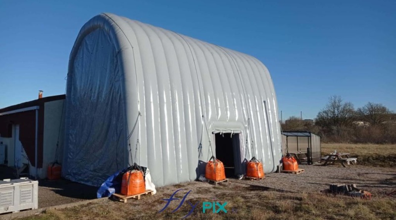 The inflatable hangar is arranged in an inverted U-shape, secured by guy lines to sand-filled ballast bags. The side access door is visible. The 0.6 mm double-skinned, padded PVC walls are very robust and strong. – All rights reserved, copyright FLYPIX.