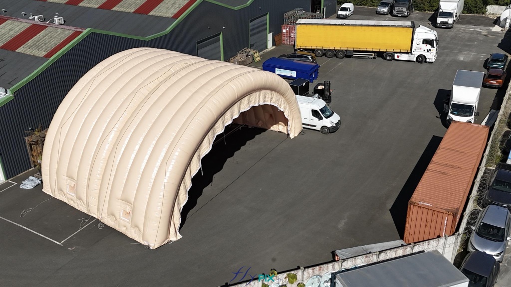 In this aerial photograph taken during an inflation and pressurization test phase prior to shipment to the client, you can see two assembled and connected half-moon tunnel modules, part of a large inflatable military hangar composed of five modules in total. Here, we have inflated the front gable module and an intermediate module. In this aerial photograph taken during an inflation and pressurization test before shipment to the client, you can see two assembled and connected half-moon tunnel modules, part of a large inflatable military hangar composed of five modules in total. Here, we have inflated the front gable module and an intermediate module. This large, wide, and tall mobile inflatable military hangar, designed for multiple uses, protects equipment and personnel from the elements during wartime. It will be deployed in military operations areas in armed conflict zones to accommodate soldiers, military personnel, paratroopers, and officers, as well as to store equipment, military vehicles, armored vehicles, tanks, aircraft, helicopters, and more. This inflatable structure, custom-made by us in our workshops in Orléans, France, is continuously inflated by several electric turbines. The inflatable shelter is entirely made of 0.45 mm double-skinned padded PVC, and features large doors, windows, and useful interfaces such as air sleeves for climate control inside the inflatable tent, and electrical cable sleeves, service doors and additional access for military personnel… – Copyright: Vivien LAÏLLE/FLYPIX, all rights reserved.