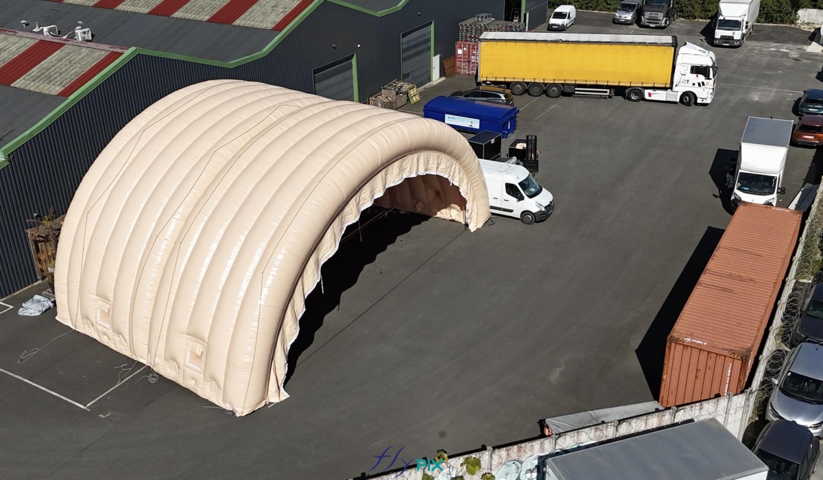 In this aerial photograph taken during an inflation and pressurization test before shipment to the client, you can see two assembled and connected half-moon tunnel modules, part of a large inflatable military hangar composed of five modules in total. Here, we have inflated the front gable module and an intermediate module. This large, wide, and tall mobile inflatable military hangar, designed for multiple uses, protects equipment and personnel from the elements during wartime. It will be deployed in military operations areas in armed conflict zones to accommodate soldiers, military personnel, paratroopers, and officers, as well as to store equipment, military vehicles, armored vehicles, tanks, aircraft, helicopters, and more. This inflatable structure, custom-made by us in our workshops in Orléans, France, is continuously inflated by several electric turbines. The inflatable shelter is entirely made of 0.45 mm double-skinned padded PVC, and features large doors, windows, and useful interfaces such as air sleeves for climate control inside the inflatable tent, and electrical cable sleeves, service doors and additional access for military personnel… – Copyright: Vivien LAÏLLE/FLYPIX, all rights reserved.
