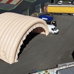 In this aerial photograph taken during an inflation and pressurization test before shipment to the client, you can see two assembled and connected half-moon tunnel modules, part of a large inflatable military hangar composed of five modules in total. Here, we have inflated the front gable module and an intermediate module. This large, wide, and tall mobile inflatable military hangar, designed for multiple uses, protects equipment and personnel from the elements during wartime. It will be deployed in military operations areas in armed conflict zones to accommodate soldiers, military personnel, paratroopers, and officers, as well as to store equipment, military vehicles, armored vehicles, tanks, aircraft, helicopters, and more. This inflatable structure, custom-made by us in our workshops in Orléans, France, is continuously inflated by several electric turbines. The inflatable shelter is entirely made of 0.45 mm double-skinned padded PVC, and features large doors, windows, and useful interfaces such as air sleeves for climate control inside the inflatable tent, and electrical cable sleeves, service doors and additional access for military personnel… – Copyright: Vivien LAÏLLE/FLYPIX, all rights reserved.