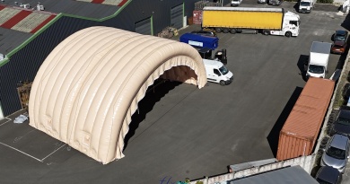 In this aerial photograph taken during an inflation and pressurization test before shipment to the client, you can see two assembled and connected half-moon tunnel modules, part of a large inflatable military hangar composed of five modules in total. Here, we have inflated the front gable module and an intermediate module. This large, wide, and tall mobile inflatable military hangar, designed for multiple uses, protects equipment and personnel from the elements during wartime. It will be deployed in military operations areas in armed conflict zones to accommodate soldiers, military personnel, paratroopers, and officers, as well as to store equipment, military vehicles, armored vehicles, tanks, aircraft, helicopters, and more. This inflatable structure, custom-made by us in our workshops in Orléans, France, is continuously inflated by several electric turbines. The inflatable shelter is entirely made of 0.45 mm double-skinned padded PVC, and features large doors, windows, and useful interfaces such as air sleeves for climate control inside the inflatable tent, and electrical cable sleeves, service doors and additional access for military personnel… – Copyright: Vivien LAÏLLE/FLYPIX, all rights reserved.
