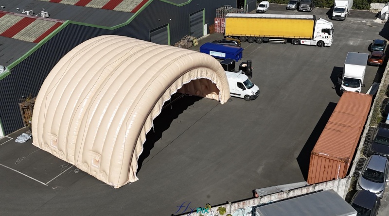 In this aerial photograph taken during an inflation and pressurization test before shipment to the client, you can see two assembled and connected half-moon tunnel modules, part of a large inflatable military hangar composed of five modules in total. Here, we have inflated the front gable module and an intermediate module. This large, wide, and tall mobile inflatable military hangar, designed for multiple uses, protects equipment and personnel from the elements during wartime. It will be deployed in military operations areas in armed conflict zones to accommodate soldiers, military personnel, paratroopers, and officers, as well as to store equipment, military vehicles, armored vehicles, tanks, aircraft, helicopters, and more. This inflatable structure, custom-made by us in our workshops in Orléans, France, is continuously inflated by several electric turbines. The inflatable shelter is entirely made of 0.45 mm double-skinned padded PVC, and features large doors, windows, and useful interfaces such as air sleeves for climate control inside the inflatable tent, and electrical cable sleeves, service doors and additional access for military personnel… – Copyright: Vivien LAÏLLE/FLYPIX, all rights reserved.