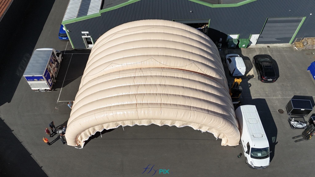 In this aerial photo, it is possible to see the type of wall, made of a solid material, as it is entirely made of 0.45 mm double-skinned padded PVC, which provides both thermal and acoustic insulation, and great strength to the entire inflatable structure. This aerial photograph reveals the type of wall, constructed from a robust material: a double-skinned, padded 0.45 mm PVC envelope. This provides both thermal and acoustic insulation, as well as exceptional strength to the entire inflatable structure. The series of interconnected, semi-circular tunnel-shaped inflatable arches creates a sturdy and durable military inflatable hangar with excellent natural buttresses, ensuring a solid base and stability on the ground. The large inflatable shelter is guyed with Lancelin professional marine ropes and sandbags to guarantee excellent wind resistance and stability. This type of large inflatable tent solution is mobile, efficient, easy to assemble and disassemble, readily transportable, and can be deployed very quickly in times of war or conflict to meet military operational needs. In wartime, this type of military hangar can serve as shelter for storing military equipment, vehicles, and machinery; for performing maintenance on machines, aircraft or helicopter engines; as a living quarters for soldiers and officers; or as an operational base or rear post, for example. The versatility of this inflatable military hangar solution can meet numerous needs in emergency situations. – Copyright: Vivien LAÏLLE/FLYPIX, all rights reserved.