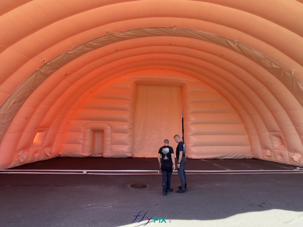 Interior view of the inflatable hangar; here, two modules are inflated, installed, and joined together. At the back, on the front gable module, a large, wide, and tall access door is visible, and to its left, a small secondary service side door. The two technicians provide a sense of the overall size of the inflatable structure, highlighting its considerable height and width. - Copyright: Vivien LAÏLLE/FLYPIX, all rights reserved. Interior view of the inflatable hangar; here, two modules are inflated, installed, and joined together. At the back, on the front gable module, a large, wide, and tall access door is visible, and to its left, a small secondary service side door. The two technicians provide a sense of the overall size of the inflatable structure, highlighting its considerable height and width. – Copyright: Vivien LAÏLLE/FLYPIX, all rights reserved.