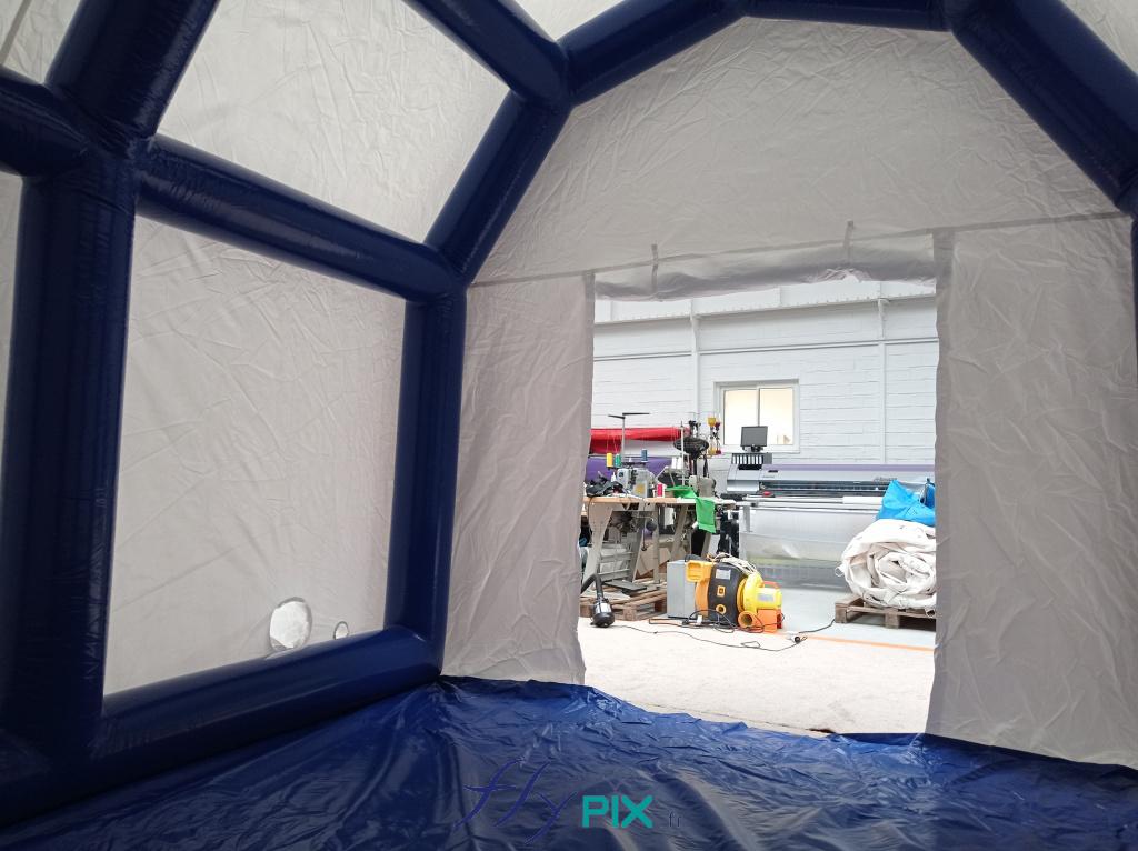 An interior view of the 4 x 3 m PMA tent : on the lower left wall , an air sleeve and an electrical cable sleeve . Below, a washable PVC groundsheet , welded to the tent . At the front, a wide gable door with wide Velcro closures . The white wall envelope is made of 0.6 mm single-layer PVC , and the airtight, sealed frame tubes are also made of 0.6 mm single-layer PVC ; the seams are welded . This inflatable medical shelter is inflated with air using an electric pump , via a non-return valve.