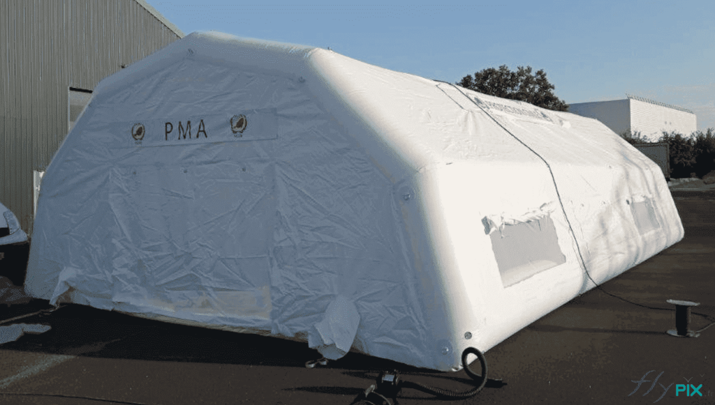 Above: a general view of the inflatable first aid post shelter, measuring L = 10 m x W = 6 m, for a usable interior area of ​​S = 60 m². This photo shows the rear gable end of the inflatable first aid post tent, with a large exit door. The roof walls feature windows and skylights with mosquito netting, transparent PVC, and removable shutters. This is a sealed-air tent, meaning the 0.6 mm single-layer PVC inflatable frame tubes are airtight and are inflated with an electric pump via a non-return valve and pressure regulator. There is an air sleeve to the left and right of the door.