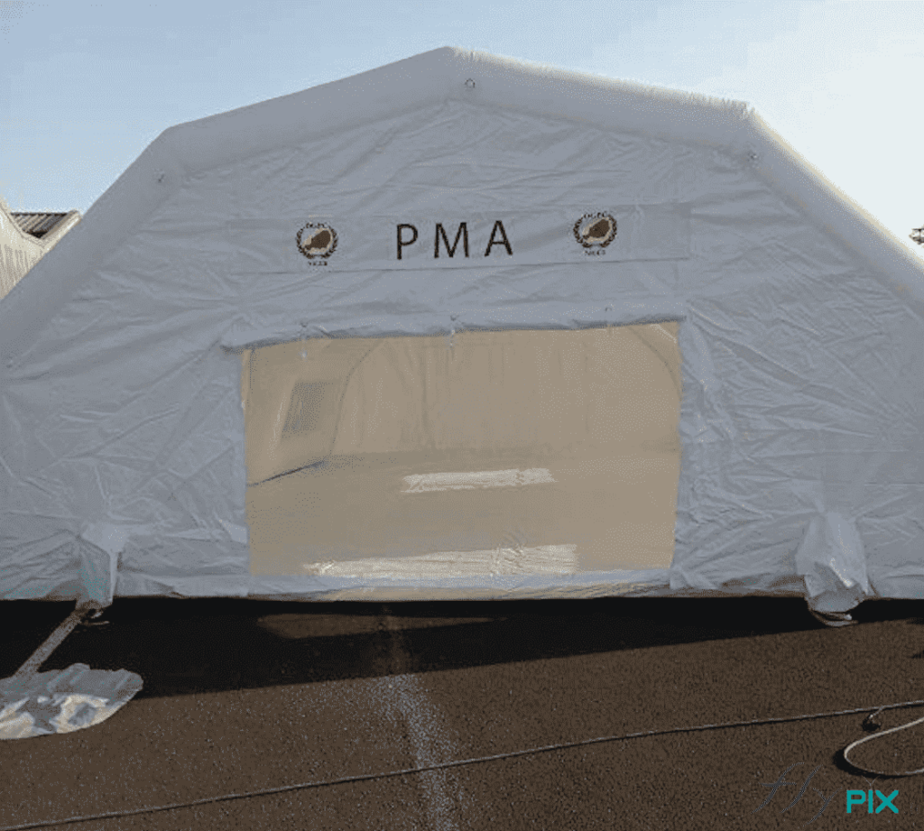A view of the rear gable end of the inflatable medical tent, with a wide exit door. To the right and left of the door, there is an air sleeve, or electrical cable conduit. The shelter has angled sides, but overall it has a tunnel shape with a keystone at a height of 2.5 m, thus providing excellent rainwater drainage in case of adverse weather.