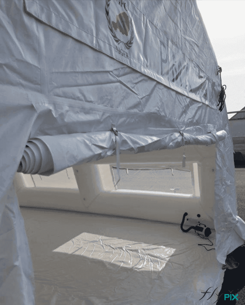Another view of the large exit door on the rear gable end of the PMA inflatable tent. The door is folded and the groundsheet is deployed. In the background, valves and a pressure regulator can be seen in operation.