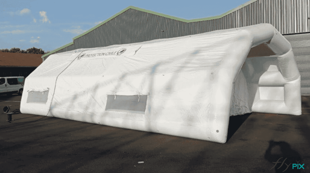 This inflatable PMA ( Advanced Medical Post ) tent, manufactured for TEDIS , is a captive air inflatable structure with internal dimensions of 10m x 6m x 2.75m (L x W x H) and a surface area of ​​60m². The inflatable shelter is entirely made of single-layer 0.6mm PVC with welded seams. It features a large, removable 60m² groundsheet. The module is inflated with air using a reversible electric pump. The kit also includes six weights and a pressure regulator. This view shows the left side with windows and the front gable end, which includes an access canopy that protects against rain and inclement weather. This type of inflatable tent takes up little space when deflated and can be deployed very quickly on any type of terrain in an emergency.